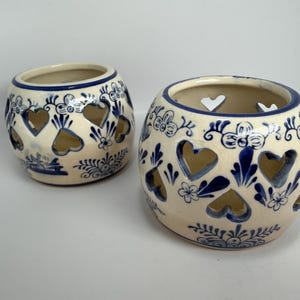Peut inclure: Deux bougeoirs ronds en céramique avec un vernis couleur crème, ornés de motifs floraux bleus peints à la main et de découpes en forme de cœur sur les côtés. Le design comprend des motifs botaniques bleus et une illustration de moulin.