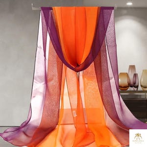 May include: A sheer, lightweight scarf featuring vertical color-blocked panels in vibrant orange and deep purple. The fabric has a translucent, airy texture and is draped over a horizontal bar, creating soft, flowing folds that hang downward.