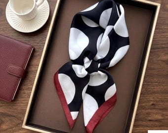 May include: A square scarf featuring a black background with large white polka dots and a solid maroon border. The scarf is folded and displayed on a dark brown tray, positioned next to a white ceramic cup and a maroon notebook.