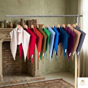 May include: A collection of triangular scarves hanging on a metal rack, displayed in a gradient of colors including cream, red, orange, mustard, green, light blue, royal blue, dark blue, purple, tan, and charcoal gray, each with a small white tag.