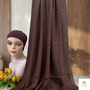 Könnte beinhalten: Ein dunkelbraunes, rechteckiges Hijab-Tuch mit dezenter Textur, das vertikal neben einem Schaufensterpuppenkopf mit passender dunkelbrauner Unterkappe drapiert ist. Die Puppe trägt einen weißen Kragen und ein kleines Sträußchen gelber Tulpen.
