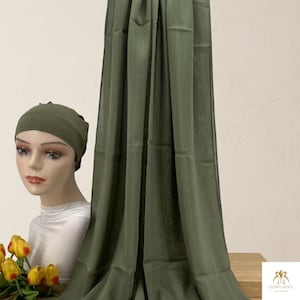 Könnte beinhalten: Ein olivgrüner Hijab, der vertikal neben einem Schaufensterpuppenkopf drapiert ist, der eine passende olivgrüne Unterkappe trägt. Die Puppe hat eine weiße Halsbedeckung. Ein kleiner Strauß gelber Tulpen befindet sich am Fuß der Puppe.