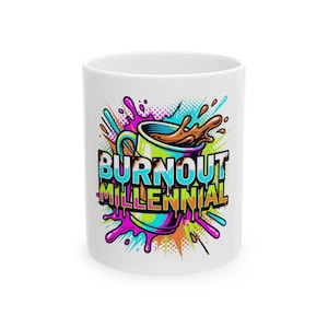 May include: A white ceramic mug featuring a colorful graphic of a coffee cup with brown liquid splashing out. The text BURNOUT MILLENNIAL is printed in bold, dripping letters over a vibrant background of pink, blue, and orange paint splatters.