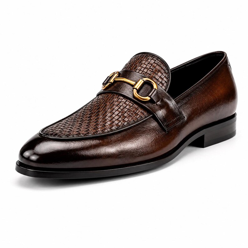 Pode incluir: Um mocassim de couro castanho escuro com uma textura entran&ccedil;ada no peito do p&eacute; e um detalhe de brid&atilde;o em metal dourado polido no topo. O sapato tem um acabamento polido suave, biqueira arredondada e um salto baixo preto com sola preta.
