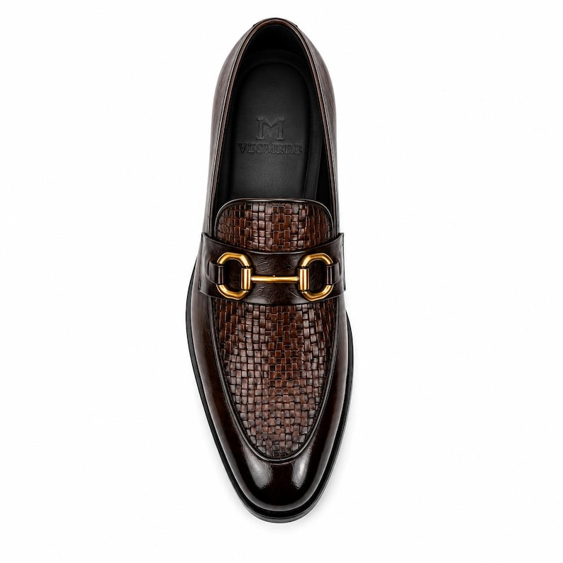 Pode incluir: Um mocassim de couro castanho escuro com textura entran&ccedil;ada no peito do p&eacute; e um detalhe de brid&atilde;o em metal dourado brilhante no topo. A palmilha interior preta apresenta um log&oacute;tipo estampado com a letra M e texto por baixo.