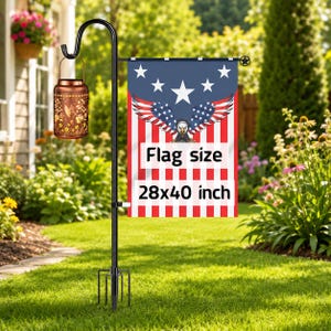 May include: A black metal garden flag stand with a four-pronged base, holding a rectangular flag featuring a bald eagle with American flag wings and the text Flag size 28x40 inch. A copper-colored cylindrical lantern hangs from the shepherd hook.