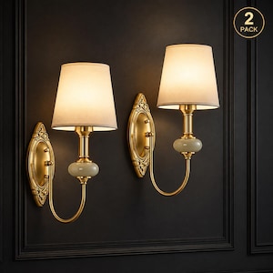 May include: A set of two wall sconces featuring gold-toned metal hardware, an ornate oval backplate, a central pale green stone accent, and a tapered off-white fabric shade. The lamps emit a warm glow and include a 2 PACK label in the corner.