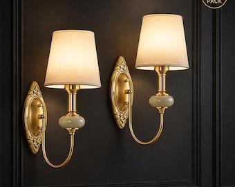 May include: A set of two wall sconces featuring gold-toned metal hardware, an ornate oval backplate, a central pale green stone accent, and a tapered off-white fabric shade. The lamps emit a warm glow and include a 2 PACK label in the corner.