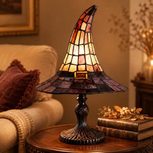May include: A stained glass table lamp shaped like a pointed hat. The lamp features a tall, curved crown with orange and cream glass panels and a wide brim made of dark purple glass. A small rectangular buckle detail sits at the base of the crown.