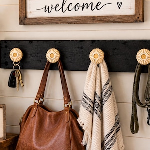 May include: A black wooden wall-mounted rack featuring four round, cream-colored, textured knobs. The rack holds a set of keys, a brown leather handbag, a cream and black striped fabric towel, and a green leash hanging from the individual knobs.