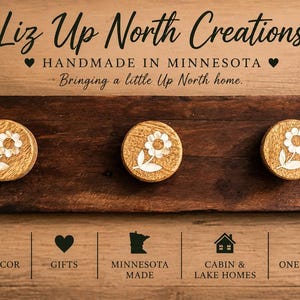 May include: A dark brown wooden wall rack featuring three circular knobs, each decorated with a white flower graphic. The text reads: Liz Up North Creations, Handmade in Minnesota, Bringing a little Up North home, Home Looks Better Up North, Rustic Decor, Functional Beauty, Minnesota Made, Small Business, Big Dreams, Home Decor, Gifts, Cabin & Lake Homes, One of a Kind, Simple Beautiful Useful.