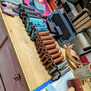 May include: A variety of leather keychains in different colors and patterns, including brown, blue, and black. The keychains are arranged in a row on a wooden surface. There are also some leather scraps and tools visible in the background.