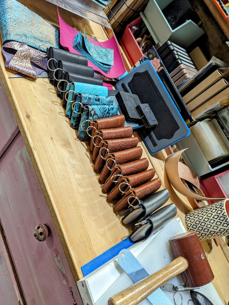 May include: A variety of leather keychains in different colors and patterns, including brown, blue, and black. The keychains are arranged in a row on a wooden surface. There are also some leather scraps and tools visible in the background.