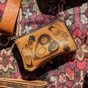 May include: A small brown leather pouch with a zipper closure. The pouch has a black and white illustration of a hand with an eye, a crescent moon, and a pentagram with the words "Hello" and "Goodbye" on it.