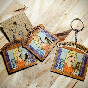 May include: Three wooden keychains with the text "MANGER BABIES" on them. Each keychain features a cartoon image of a woman with blonde hair holding a cat and a puppet. The keychains are on a wooden surface.