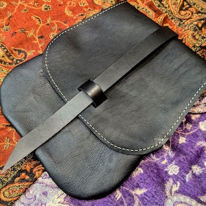 May include: A black leather pouch with a flap closure and a long strap. The pouch has a light brown stitch detail around the edges.