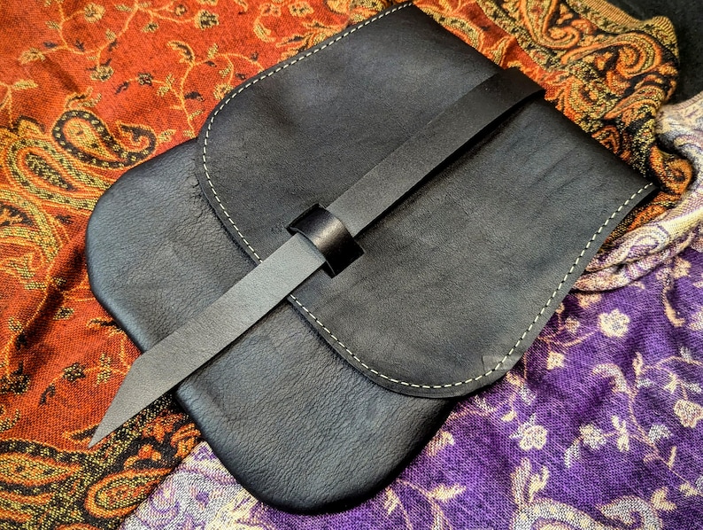 May include: A black leather pouch with a flap closure and a long strap. The pouch has a light brown stitch detail around the edges.