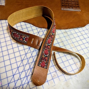 Op de afbeelding: Een bruine leren gitaarband met een centraal geweven stoffen inzetstuk met een gedetailleerd bloemmotief in tinten rood, blauw en goud. De band is voorzien van verstevigde leren uiteinden met voorgesneden gaten voor bevestiging aan een instrument.