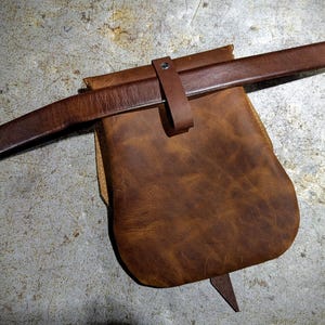 May include: A brown leather pouch with a brown leather strap. The pouch has a flap that closes with a buckle.