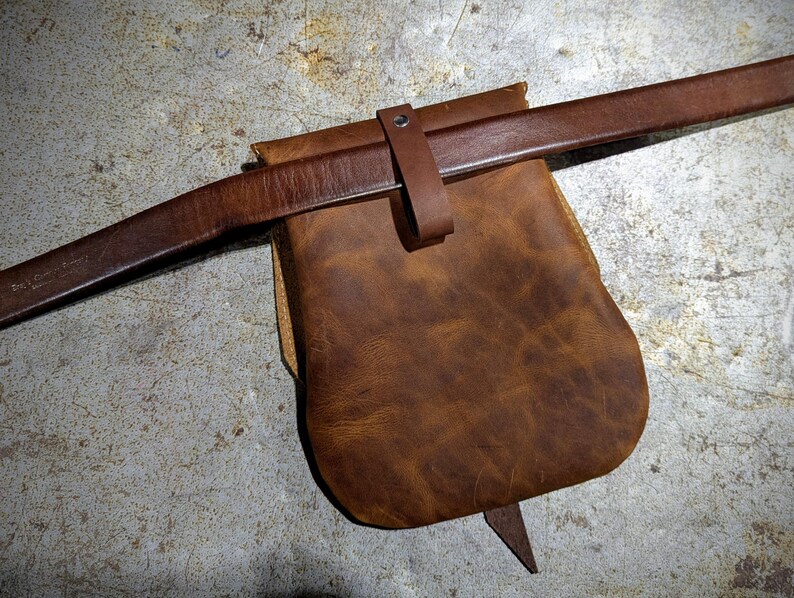 May include: A brown leather pouch with a brown leather strap. The pouch has a flap that closes with a buckle.