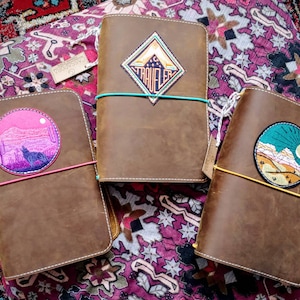 Pode incluir: Três diários de couro castanho com diferentes patches bordados. Os patches apresentam uma cordilheira com uma lua crescente, um lobo a uivar numa paisagem desértica e uma cordilheira com um sol.