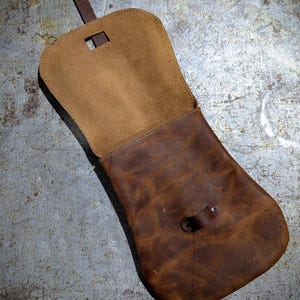 May include: A brown leather bag with a flap closure and a strap. The bag is open and the inside is visible. The bag is made of a soft, supple leather and has a rustic look.