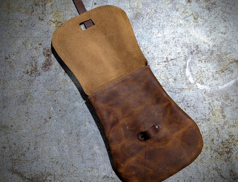 May include: A brown leather bag with a flap closure and a strap. The bag is open and the inside is visible. The bag is made of a soft, supple leather and has a rustic look.