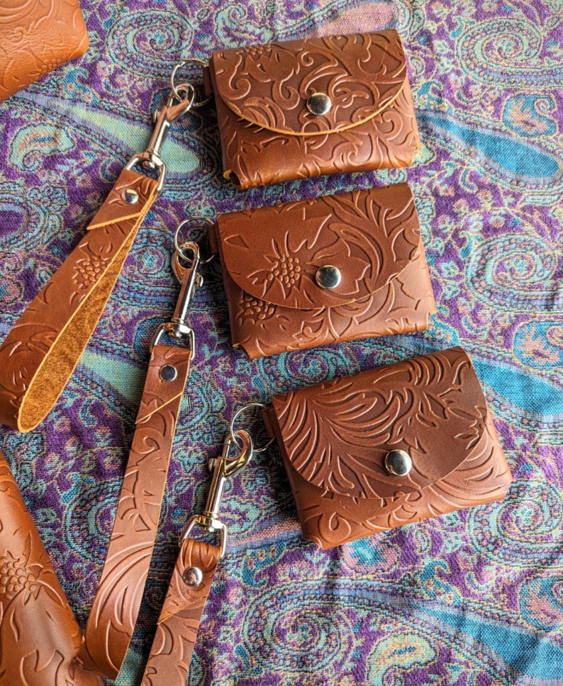 May include: Three brown leather keychains with a floral embossed design. Each keychain has a metal clasp and a small pocket.