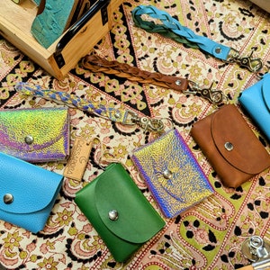 May include: Assortment of small leather wallets with iridescent, blue, green, and brown finishes. Each wallet has a snap closure and a metal ring for attaching keys. The wallets are displayed on a patterned fabric surface.
