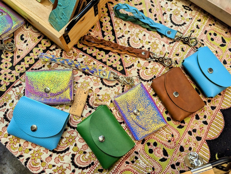 May include: Assortment of small leather wallets with iridescent, blue, green, and brown finishes. Each wallet has a snap closure and a metal ring for attaching keys. The wallets are displayed on a patterned fabric surface.