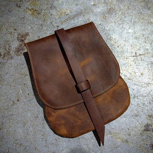 May include: A brown leather satchel with a long strap. The bag has a flap with a buckle closure and is made of distressed leather.