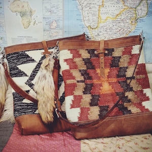 May include: Two leather tote bags with colorful woven fabric panels. The bag on the left has a black and white geometric pattern. The bag on the right has a red, orange, brown, and white geometric pattern. Both bags have brown leather straps and handles.