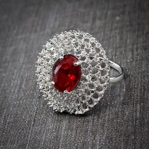 Puede incluir: Un anillo de tono plateado con una gran piedra central ovalada de color rojo intenso. La piedra está rodeada por un engaste circular ancho de estilo filigrana, incrustado con numerosas piedras pequeñas, claras y brillantes en un patrón de red.