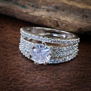 Può includere: Un anello color argento con una grande pietra centrale rotonda, trasparente e sfaccettata, fissata da sei griffe. La fascia è composta da tre file distinte, con le due file esterne incastonate di piccole pietre trasparenti e scintillanti.