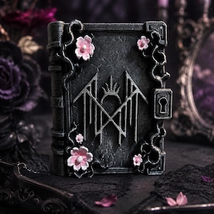 May include: A dark gray, rectangular book-shaped box with a textured, stone-like finish. The cover features a central silver geometric emblem, a keyhole on the right side, and sculpted vine details adorned with small, light pink floral accents.