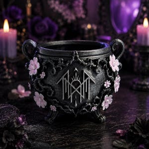 May include: A black cauldron with two handles and three feet, featuring a silver-toned geometric emblem on the front. The exterior is decorated with sculpted vines and small, light pink five-petaled flowers arranged around the central symbol.