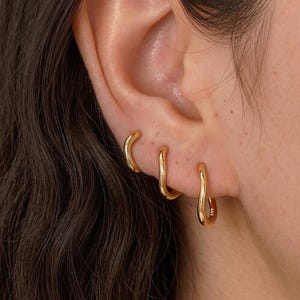 Könnte beinhalten: Drei goldfarbene Creolen mit einer glatten, wellenförmigen und unregelmäßigen Silhouette. Jeder Ohrring hat eine polierte Oberfläche und eine geschwungene, organische Form, die in einer Reihe am Ohrläppchen angeordnet ist.