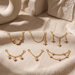 Könnte beinhalten: Sechs goldfarbene Metall-Schmuckverbindungsketten mit verschiedenen Designs, darunter ein Halbmond, vierzackige Sterne und klare, runde Zirkonia-Steine. Jedes Stück hat kleine kreisförmige Ösen an beiden Enden zur Befestigung.