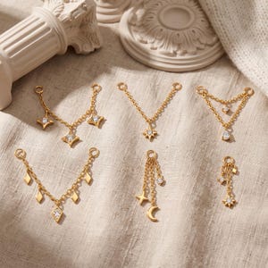May include: A collection of six gold-toned metal jewellery connectors featuring delicate chains and small clear cubic zirconia stones. The designs include star, moon, and geometric shapes, each equipped with small circular loops for attachment.