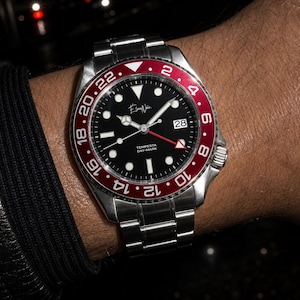 May include: A stainless steel wristwatch with a black dial, luminous hour markers, and a date window at the three o'clock position. The watch features a bidirectional rotating bezel in red and black with white numerals and a red GMT hand.