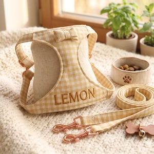 May include: A light yellow and white gingham pet harness featuring a small bow at the top and the name LEMON embroidered in gold thread. The set includes a matching gingham lead with rose gold metal clips and a small bone-shaped tag.