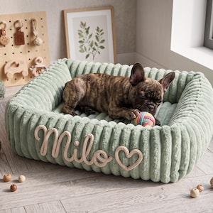 May include: A rectangular sage green pet bed with a ribbed, plush texture. The front features the name Milo and a heart shape in beige rope lettering. A brindle French Bulldog is lying inside the bed, playing with a small, multicolored yarn ball.