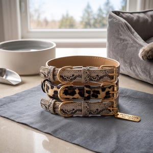 May include: A stack of three pet collars with gold-toned metal buckles. The top and bottom collars feature a grey and black snakeskin pattern, while the middle collar has a brown and black leopard print. A gold tag reads PETCOLLARS & CO. - ETSY.