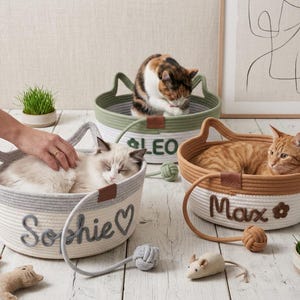 Puede incluir: Tres camas redondas de cuerda tejida con asas en forma de orejas de gato. Una cama blanca y gris con el nombre Sophie, una blanca y verde con Leo y una blanca y marrón con Max. Cada cama incluye un juguete de cuerda y un ratón de peluche.