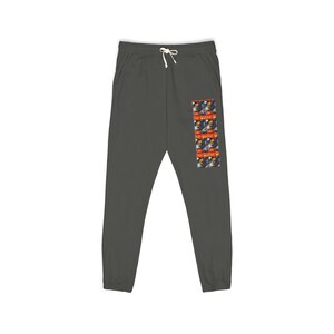 May include: Dark gray sweatpants featuring a white drawstring at the elastic waistband. A vertical rectangular graphic print on the right leg displays a repeating pattern of a comic book style illustration with the text MYSTIC in yellow letters.