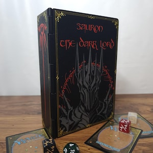 Puede incluir: Una caja de cartas rectangular negra con un gráfico de Sauron en gris y rojo, con el texto Sauron The Dark Lord en letras rojas. La caja está decorada con bordes dorados e incluye dados de juego y cartas de juego a su alrededor.
