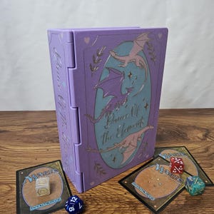 May include: A lavender rectangular deck box featuring a central oval with three flying dragons and the text Dance Of The Elements. The box is accompanied by three Magic The Gathering cards and three multi-sided dice in red, blue, and green.