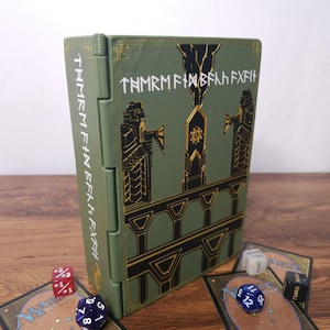 May include: A rectangular olive green storage box with a hinged side, featuring black and gold architectural illustrations and white runic text. The box is surrounded by several game cards and multi-sided dice in red, blue, white, and black colors.