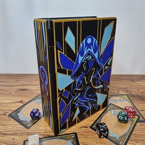 Puede incluir: Una caja de cartas rectangular negra con bordes dorados y un gráfico de una figura encapuchada azul. La caja cuenta con un lomo articulado y un diseño estilizado de una persona lanzando magia. Varios dados y cartas rodean la caja.
