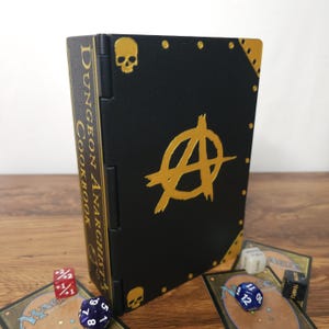 May include: A black rectangular storage box with gold accents, featuring a skull at the top and bottom, a large anarchy symbol in the center, and the text "Dungeon Anarchist's Cookbook" printed vertically on the spine, accompanied by game dice.